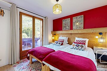 A cozy bedroom with two single beds, a vibrant red accent wall, wooden headboard, and a patio door opening to a green mountain view.