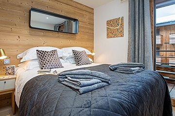 Cozy chalet bedroom with a wood-paneled wall, a mirror above the bed, a dark grey quilt, folded towels, and a balcony on the right.