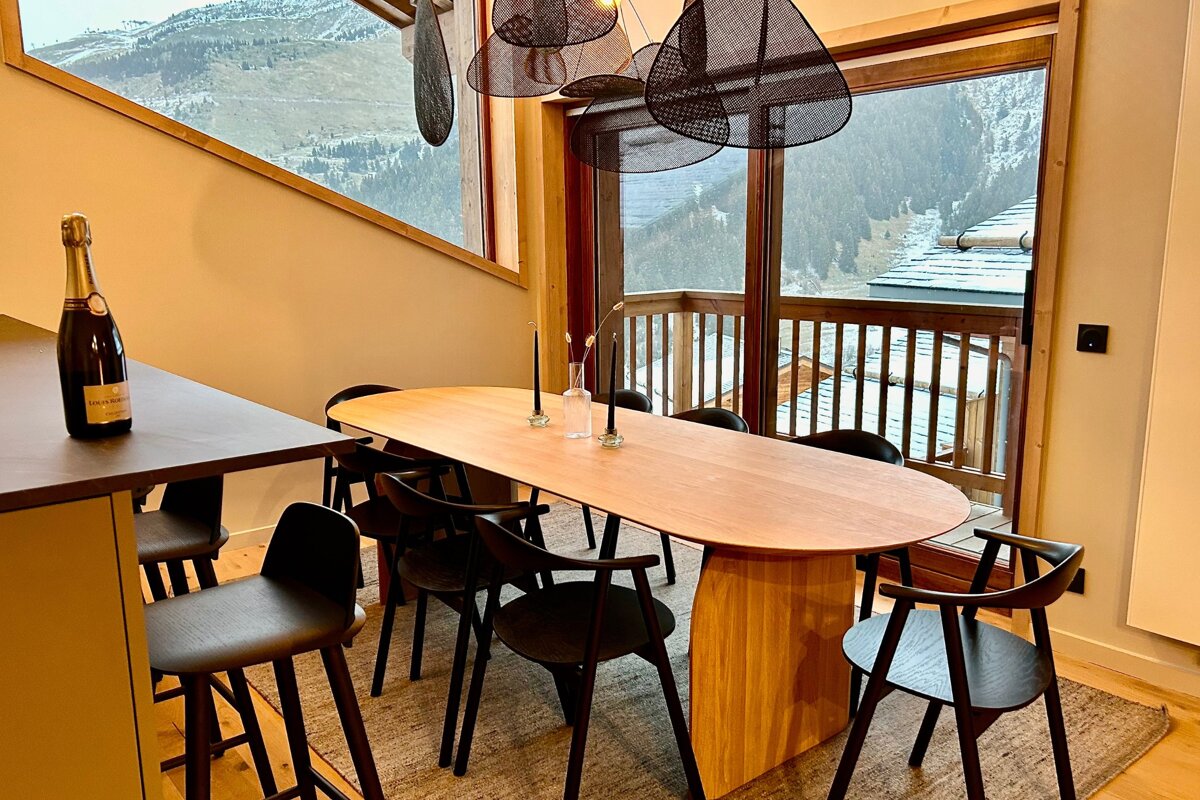 A modern dining area with a long wooden table, black chairs, and a champagne bottle, overlooking snowy mountains from large windows.