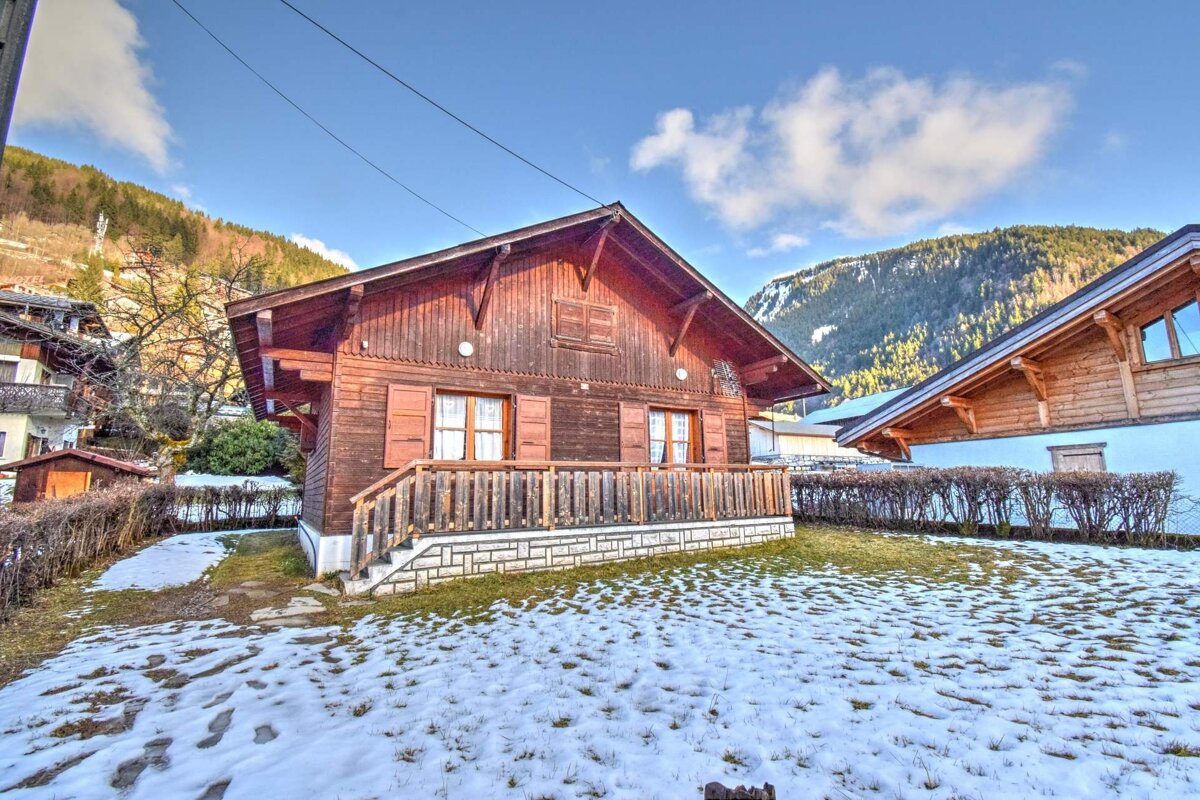A brown wooden chalet with a balcony, snow on the ground, and forested mountains under a clear blue sky.