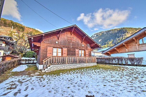 A wooden mountain chalet with a balcony, set on a snowy lawn with mountains in the background.