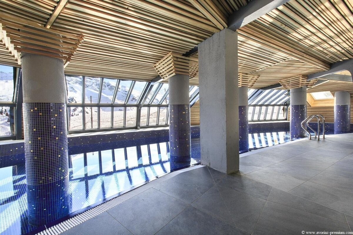 Indoor pool featuring blue tiled pillars, a slatted wood ceiling, and large windows looking out at snowy mountains.