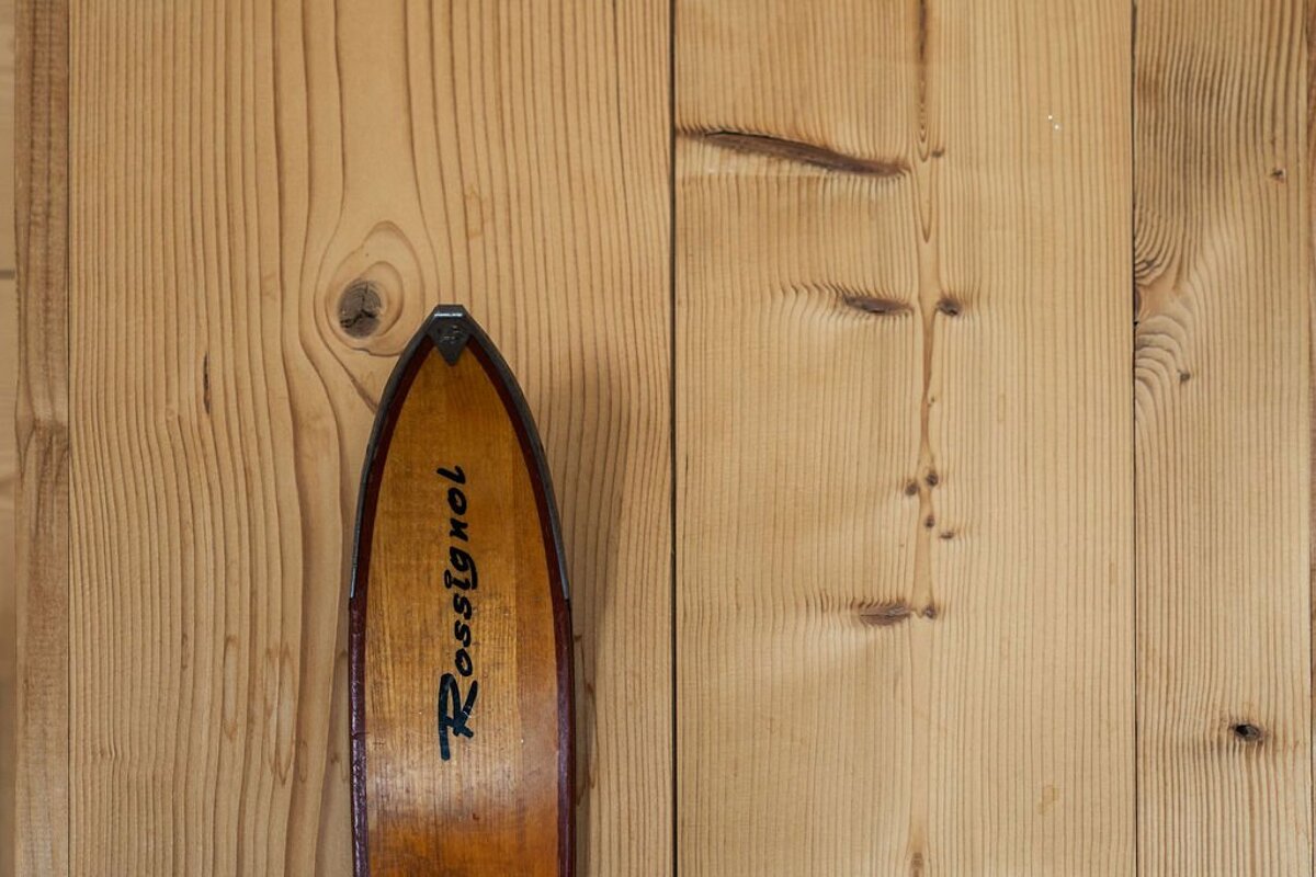 A vintage wooden Rossignol ski with a metal tip leans against a light wood plank wall with prominent grain and knots.