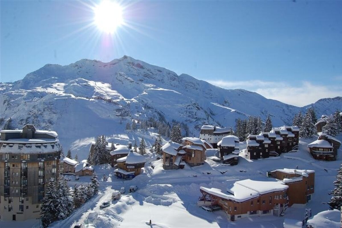 Bright sun over a snowy mountain resort village. Buildings and pine trees are blanketed in snow, with ski slopes visible on the distant peaks.