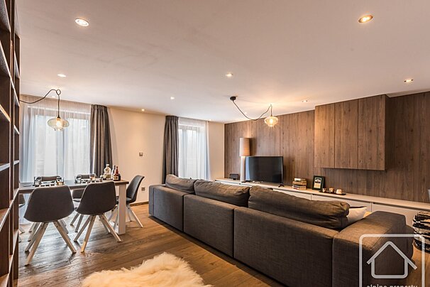 A modern open-plan living and dining room with a long grey sofa, wooden flooring, and a dining table. Features a wooden accent wall with a TV.