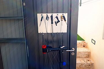 Various tools and a bike repair stand hang on a dark door. Stone steps and artificial grass are to the right, with a shed partially visible left.