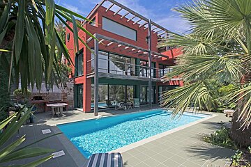 A modern red house with large windows, a swimming pool, and lush tropical foliage like palm trees, all under a bright blue sky.