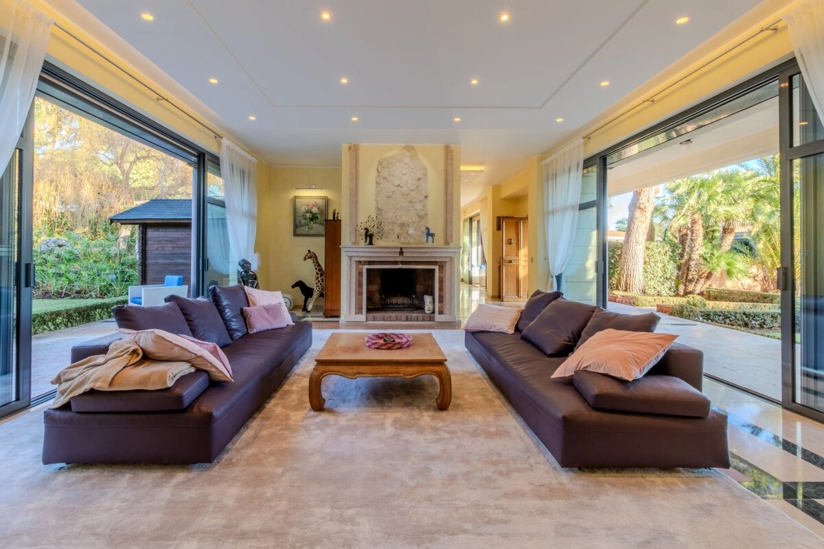 Spacious, sunlit living room with two dark sofas, a coffee table, a fireplace, and large glass doors opening to a lush garden.