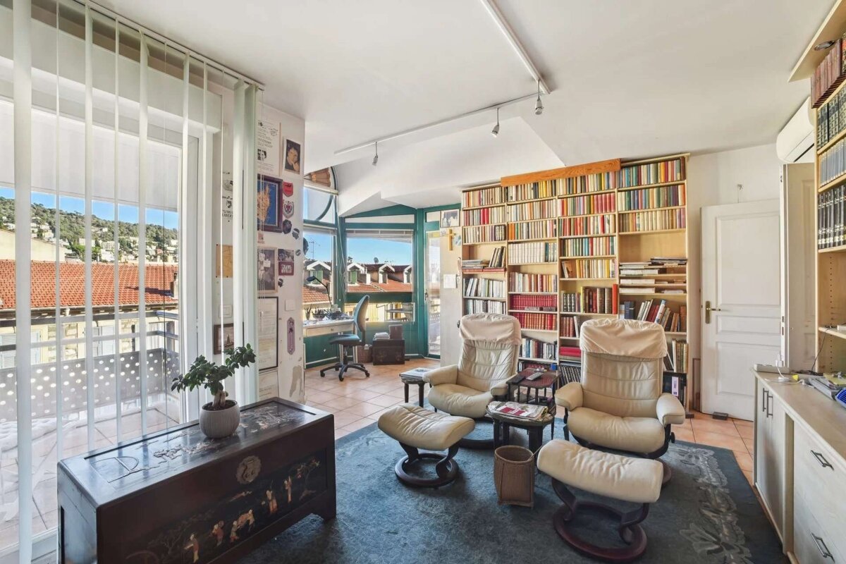 A cozy, book-filled study with two recliners, a desk by a window, and a balcony offering views of red-roofed buildings and hills.