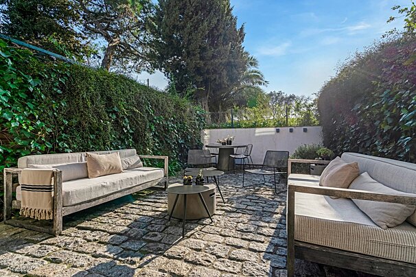 Sunny cobblestone patio with comfortable wooden sofas, lush ivy walls, and a small dining set under a blue sky.