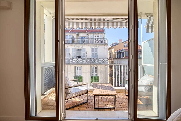 View through open doors to a sunny balcony with outdoor furniture and a striped awning, overlooking charming city buildings.