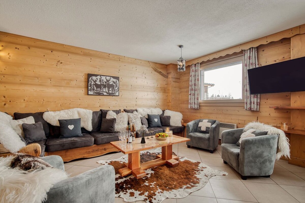 Cozy, wood-paneled chalet living room with a large sofa, armchairs, coffee table, and a cowhide rug on tiled floors. Rustic decor and deer art.