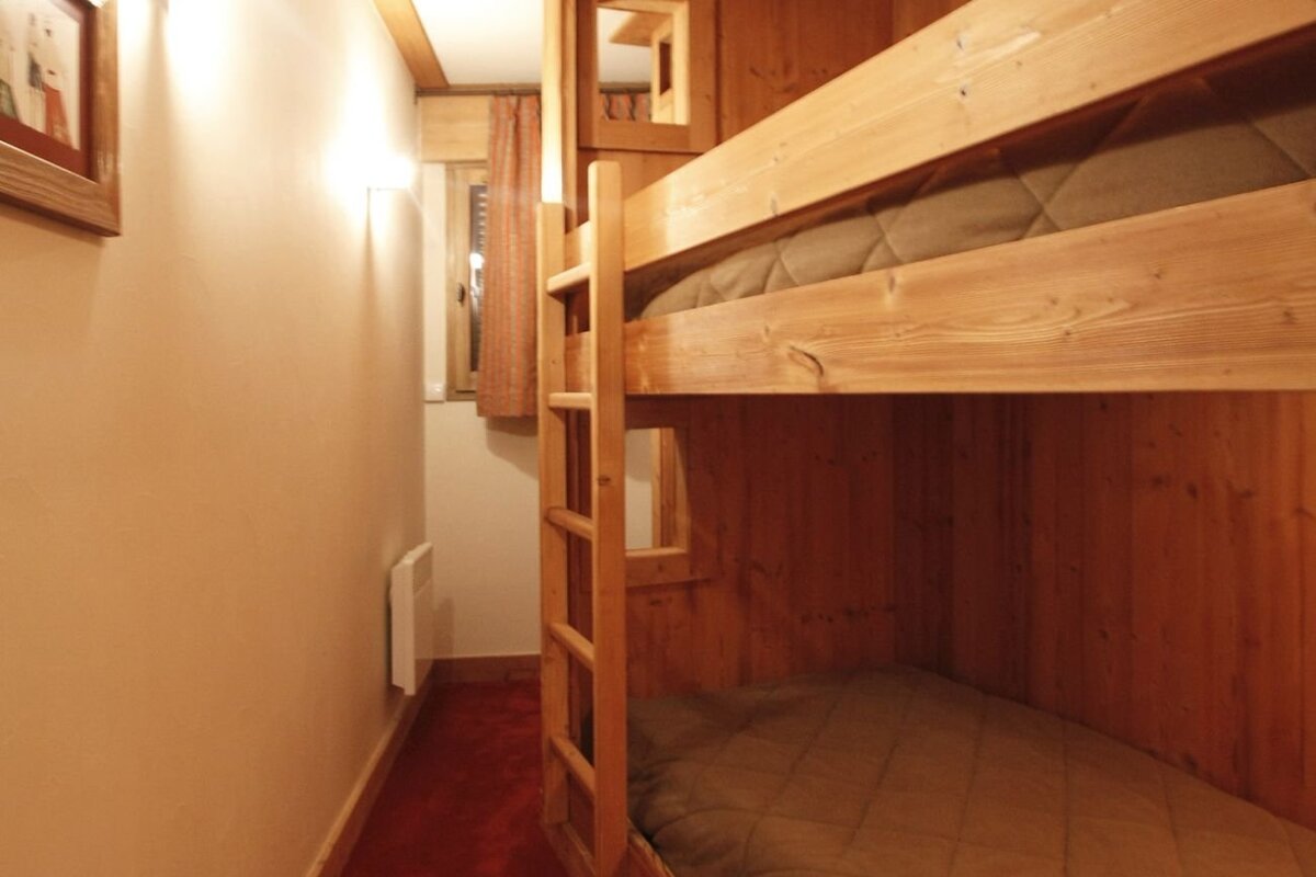 A narrow room with a rustic wooden bunk bed, ladder, quilted mattresses, red carpet, and warm lighting.