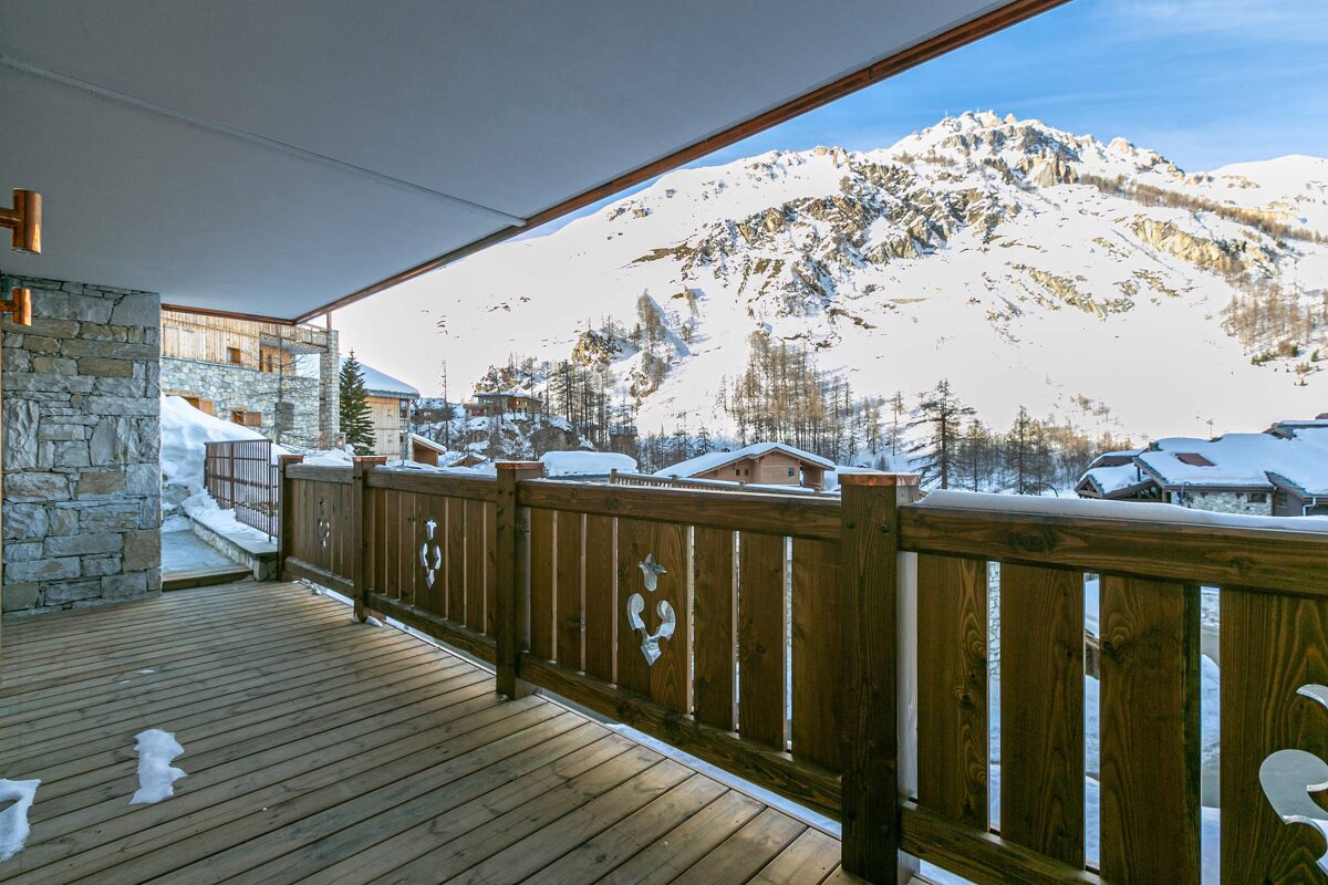 A wooden deck/balcony with carved railings offers a stunning view of snow-covered mountains and a picturesque winter village under a bright sky.