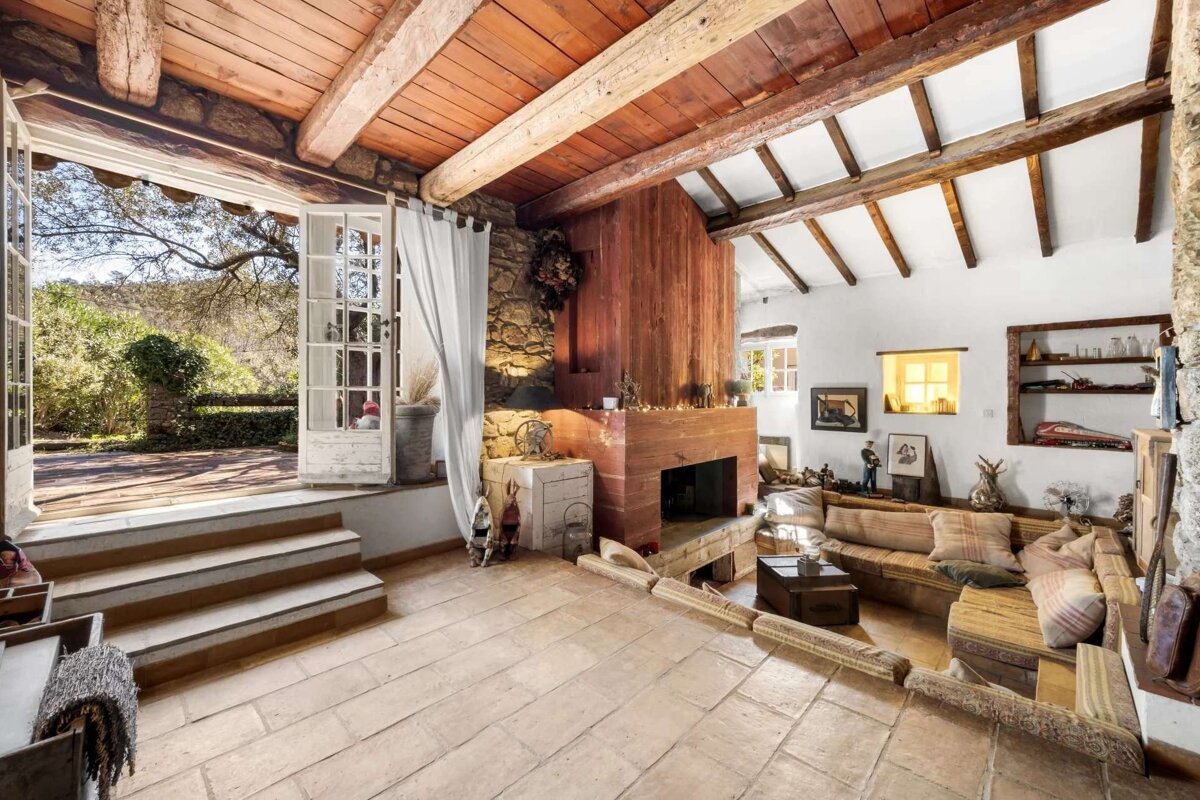 Rustic living room with a large stone/wood fireplace, beamed ceiling, tiled floor, and a sunken seating area. French doors open to a sunny patio.