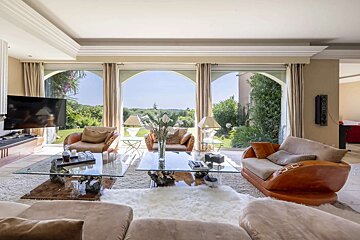 An elegant living room with tan sofas, glass tables, and expansive arched windows looking out onto a vibrant green landscape.
