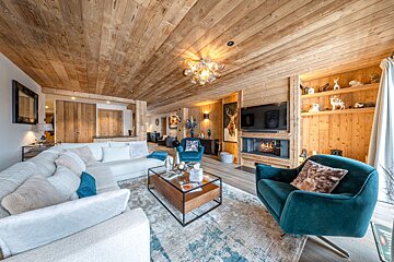 Luxurious, wood-paneled chalet living room with a white sofa, teal chairs, fireplace, and deer decor. Cozy, rustic modern style.