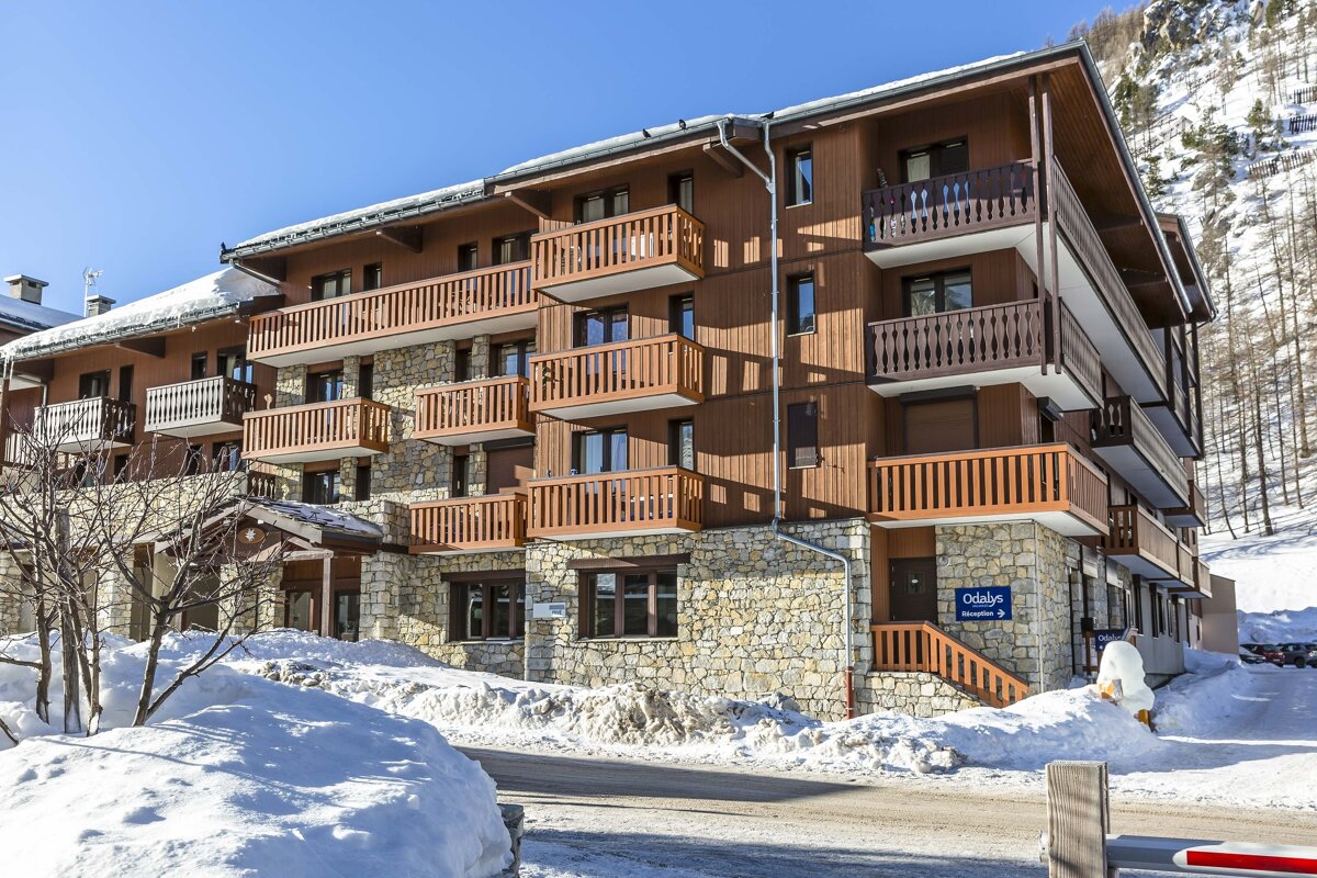 A multi-story building with wood and stone facades and balconies, nestled in a snowy, mountainous landscape under a clear blue sky.