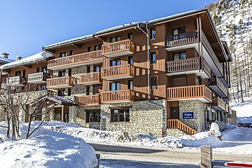 A multi-story building with wood and stone facades and balconies, nestled in a snowy, mountainous landscape under a clear blue sky.