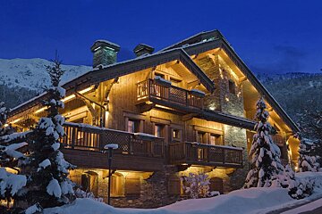 A luxurious wooden chalet glows warmly at twilight, surrounded by snow-covered trees and distant mountains, creating a cozy winter scene.