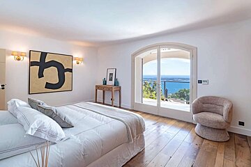 A serene white bedroom features a large bed, abstract art, and an arched door opening to a stunning blue ocean view from a balcony.