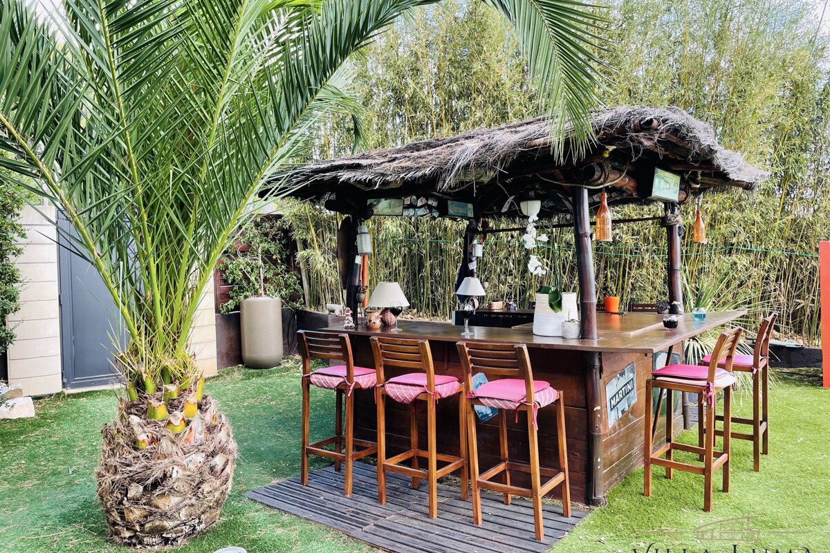 An outdoor tiki bar with a thatched roof, bar stools, and a large palm tree in a lush green garden with a bamboo backdrop.