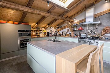 A spacious, modern kitchen with sage cabinets, a large island, and a full pantry, nestled under a rustic wooden-beamed attic ceiling.
