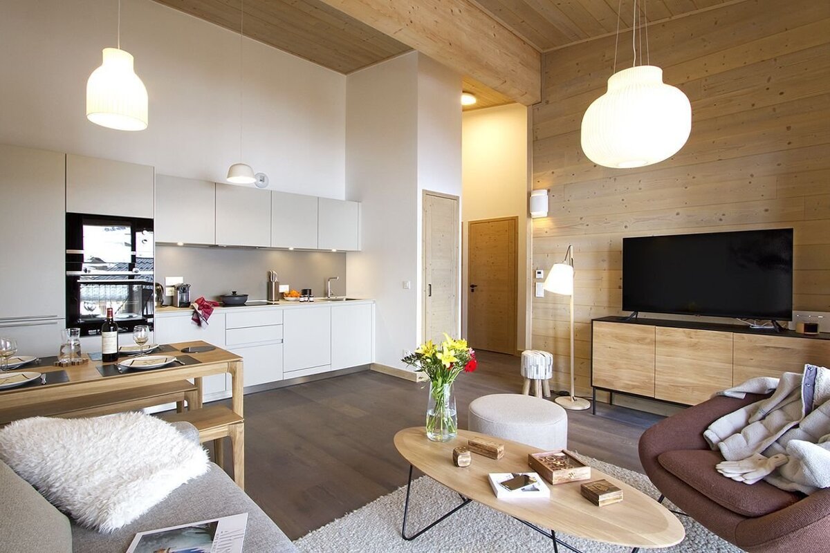 A bright, modern open-plan living area with a white kitchen, wooden dining table, TV lounge, and cozy accents, featuring light wood details.