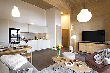 A bright, modern open-plan living area with a white kitchen, wooden dining table, TV lounge, and cozy accents, featuring light wood details.