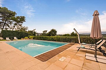 An inviting outdoor swimming pool with a tiled patio, lounge chairs, and a closed umbrella. Lush green hedges and trees frame a distant landscape under a blue sky.