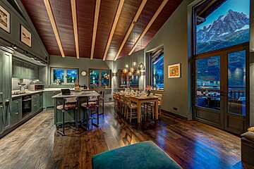 A chic, open-plan kitchen and dining area featuring green cabinetry, rich wood accents, and expansive windows framing a breathtaking mountain view. Warm chalet style.