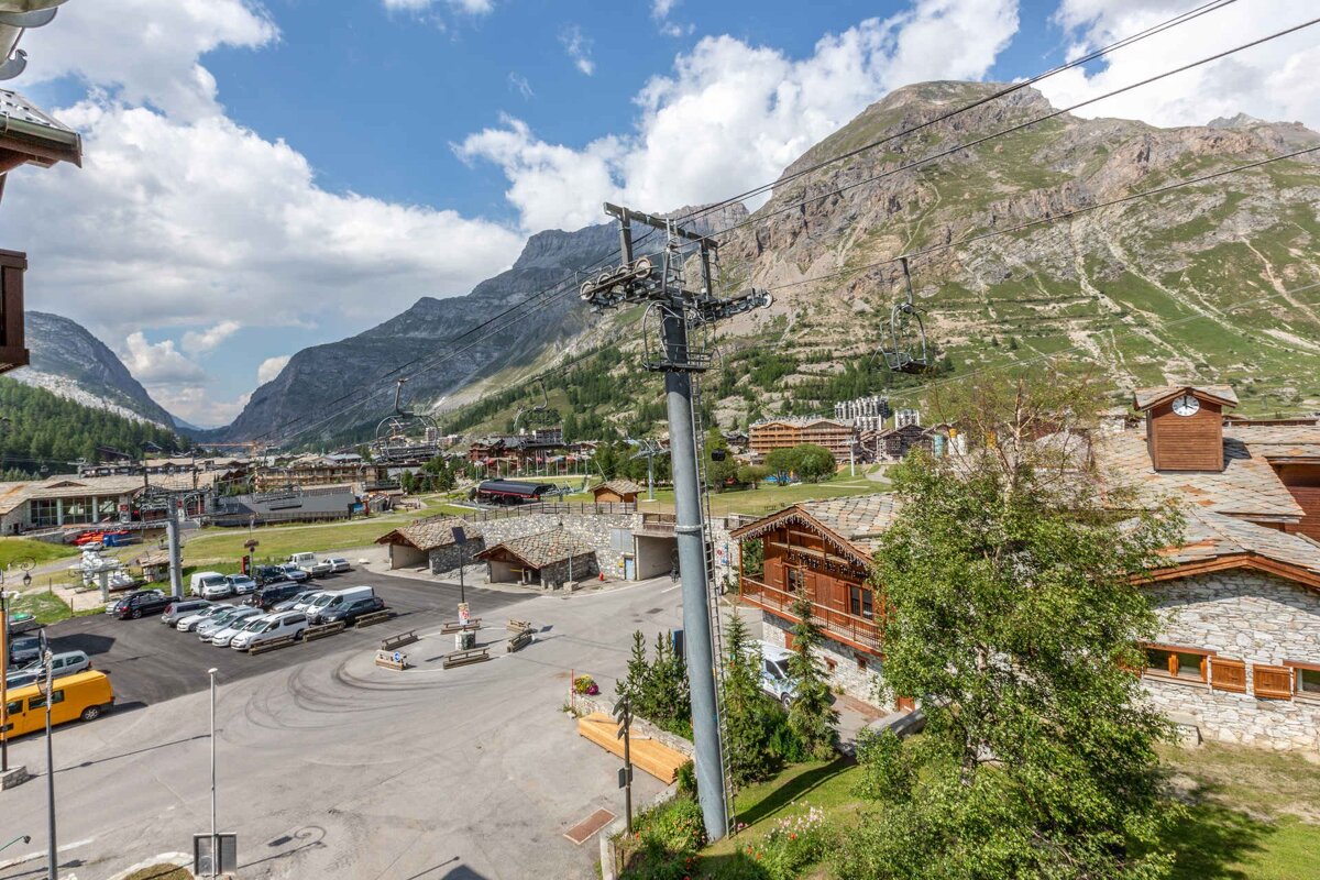 Scenic summer view of an alpine resort with stone chalets, a ski lift, and grassy mountains under a blue sky.