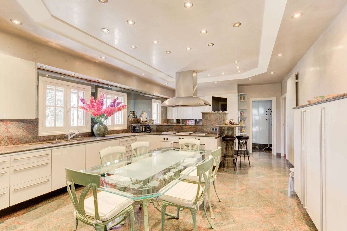 A spacious, modern kitchen and dining area with a glass table, light green chairs, white cabinets, marble countertops, and recessed lighting.