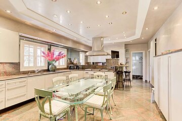 A spacious, modern kitchen and dining area with a glass table, light green chairs, white cabinets, marble countertops, and recessed lighting.