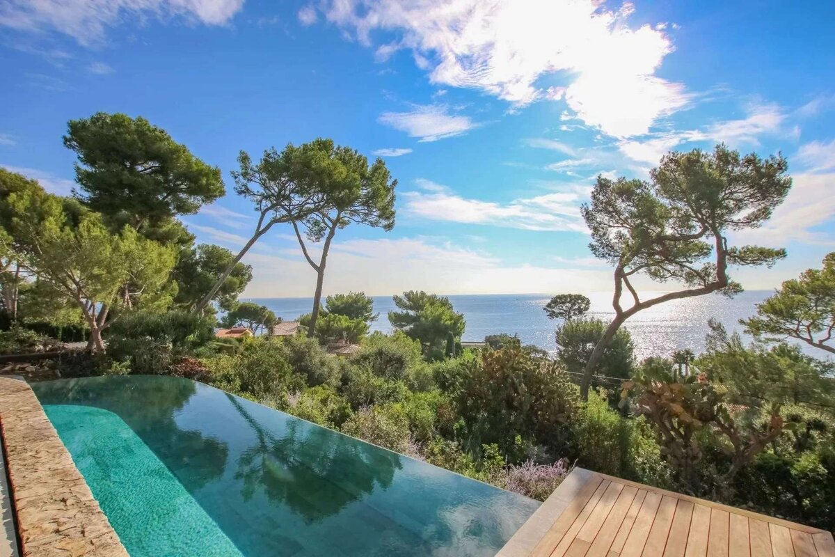 An infinity pool offers a stunning view of a sparkling blue ocean, framed by lush green trees under a bright, cloudy sky.