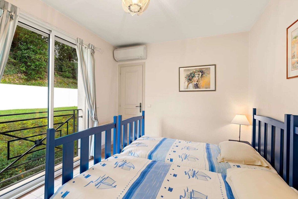 A bright bedroom with two beds featuring blue nautical bedding and headboards. A large sliding glass door opens to a balcony overlooking lush greenery.