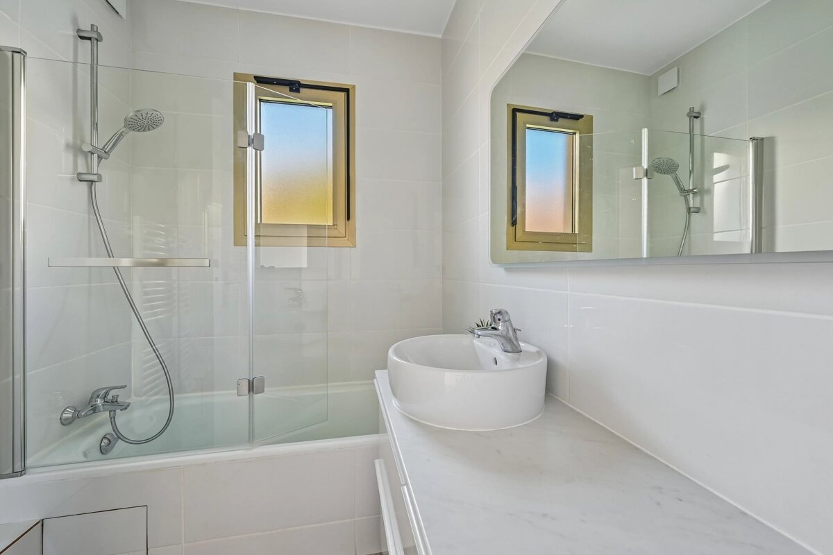 A modern white bathroom with a glass-enclosed shower/tub combo, a round vessel sink, and a large mirror reflecting the space.