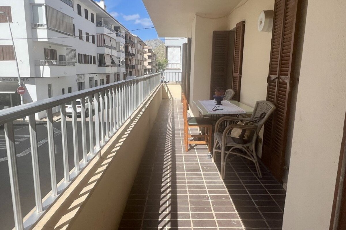 A sunlit long balcony with a table and chairs, featuring strong striped shadows and overlooking a street lined with white buildings.