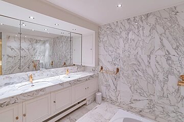 Luxurious bathroom featuring white marble walls and countertops, a double vanity with gold faucets, and a large mirror.