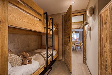 Rustic wood-paneled room with wooden bunk beds, a teddy bear on the bottom. A partially open door reveals a bright dining area.