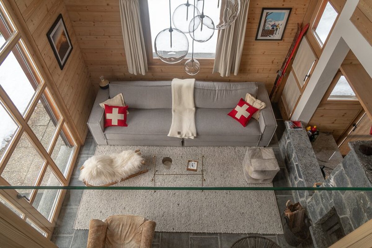 Cozy cabin living room seen from above, featuring a grey sofa with Swiss cross pillows, glass pendant lights, and rustic wood walls.