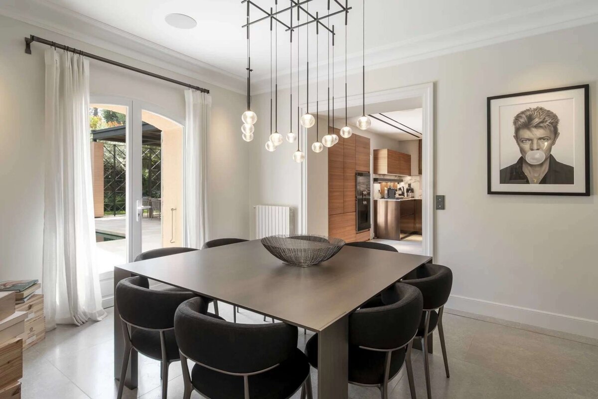 A modern dining room with a square table, black chairs, a geometric chandelier, and a David Bowie print. A patio is seen through the window, and a kitchen through an archway.