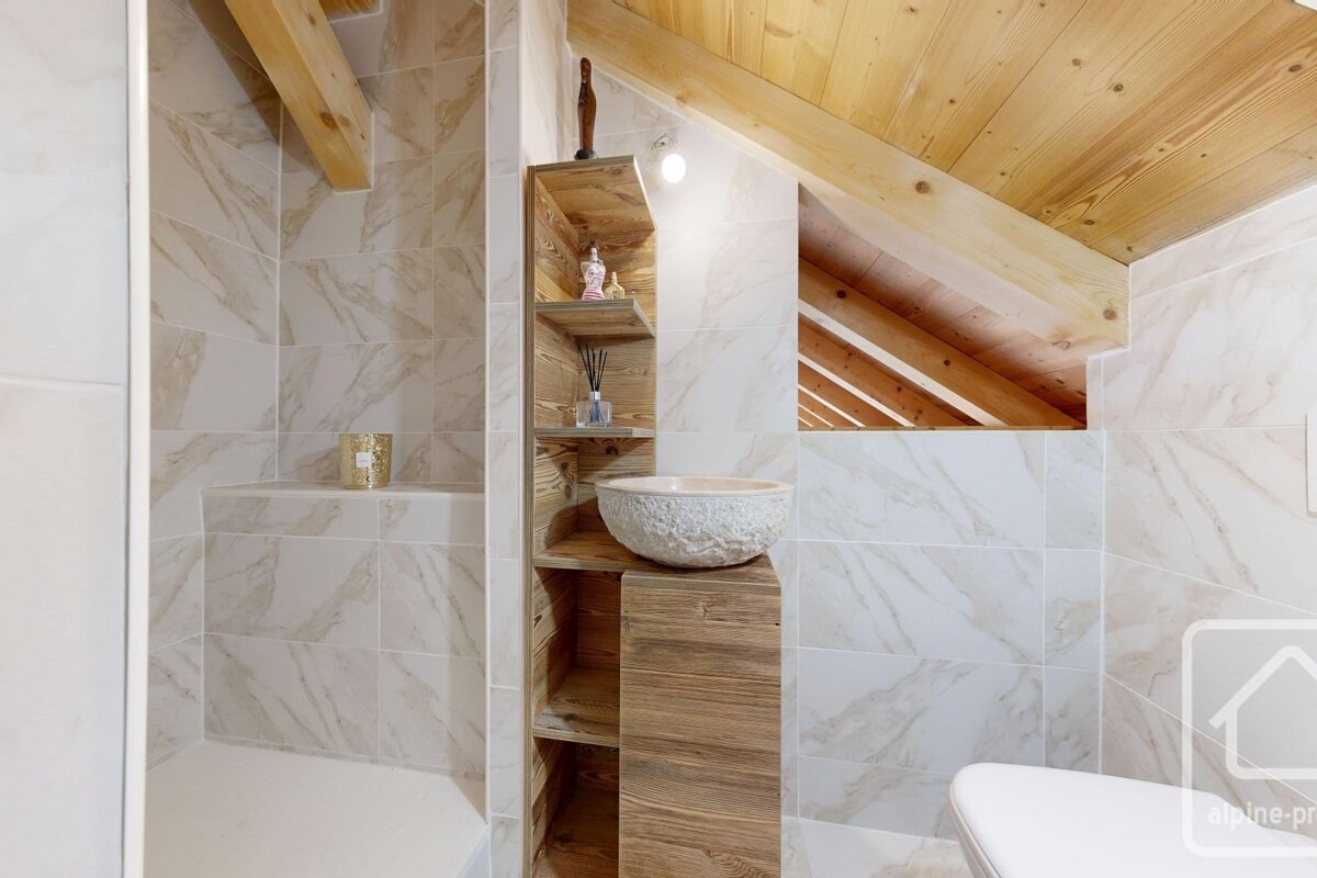 Rustic modern bathroom with marble-look tiled walls, wooden ceiling beams, a natural wood vanity with shelving, and a textured stone sink.