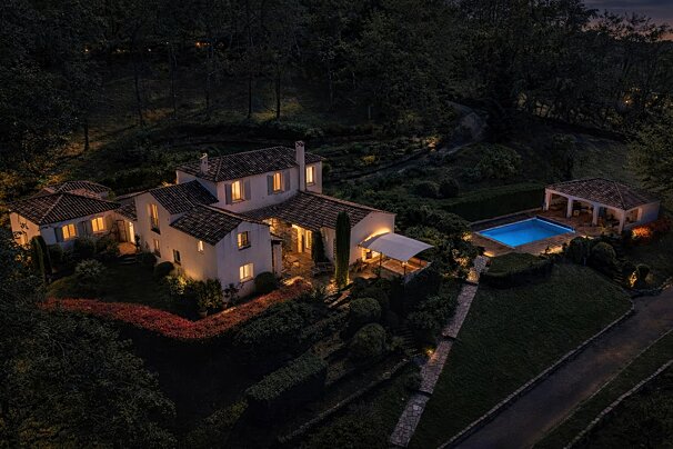 Aerial twilight view of a secluded luxury villa with glowing windows, manicured gardens, and a bright blue swimming pool.
