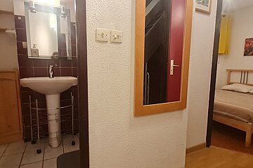 A view into a bathroom with a sink and mirror, a hallway with a mirror reflecting a red door and stairs, and a bedroom entrance with a bed.