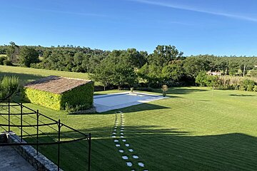 A lush green lawn with a pool, ivy-covered shed, and stepping stone path, backed by trees under a clear blue sky.