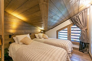 Cozy rustic attic bedroom featuring two single beds with warm knitted blankets under a slanted wooden ceiling.