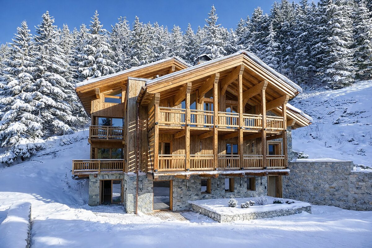 A beautiful multi-story wooden chalet with stone base is nestled in deep snow, surrounded by a dense, snow-covered pine forest under a clear sky.
