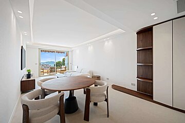 A bright, modern living room with white walls, a round wooden dining set, and a balcony offering picturesque sea and mountain views.