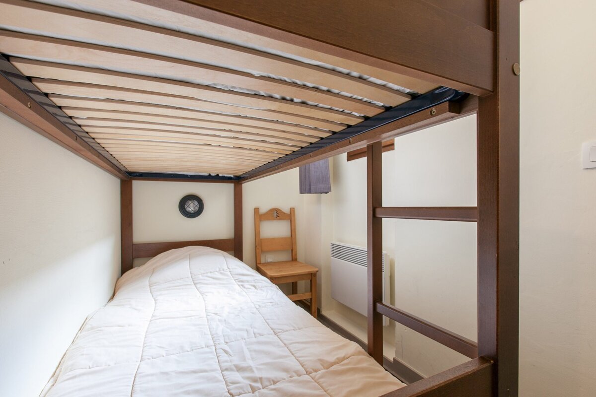 View from inside a cozy bottom bunk bed, with a white comforter, wooden frame, small chair, and wall heater visible.